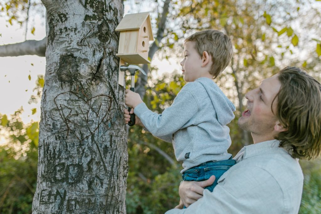 A joyful father assists his son in hanging a birdhouse on a tree, fostering a love for nature.